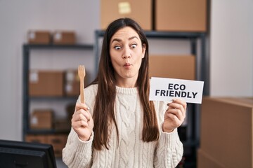 Poster - Young brunette woman working at eco friendly ecommerce making fish face with mouth and squinting eyes, crazy and comical.