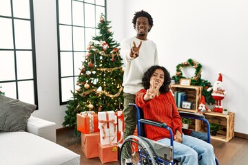 Sticker - Young interracial couple with woman sitting on wheelchair by christmas tree smiling looking to the camera showing fingers doing victory sign. number two.