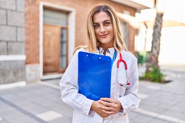 Wall Mural - Young blonde woman wearing doctor uniform holding checklist at street