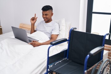 Wall Mural - Young hispanic man lying on the bed, using wheelchair smiling happy pointing with hand and finger to the side