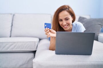 Poster - Young woman using laptop and credit card lying on sofa at home