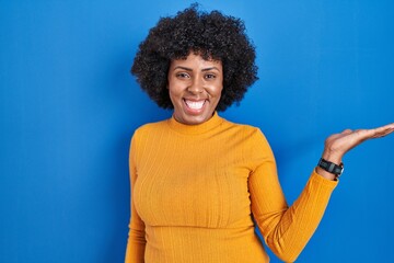 Sticker - Black woman with curly hair standing over blue background smiling cheerful presenting and pointing with palm of hand looking at the camera.