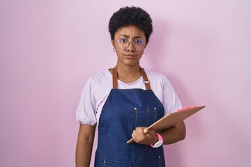 Canvas Print - Young african american woman wearing professional waitress apron holding clipboard skeptic and nervous, frowning upset because of problem. negative person.