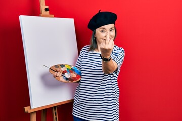 Poster - Middle age hispanic woman standing drawing with palette by painter easel stand showing middle finger, impolite and rude fuck off expression