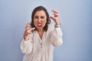 Sticker - Middle age hispanic woman standing over blue background shouting frustrated with rage, hands trying to strangle, yelling mad