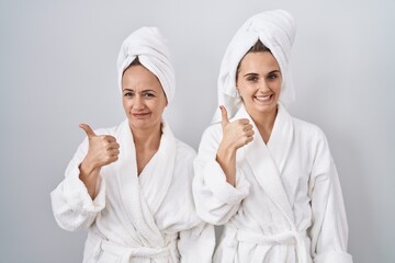 Middle age woman and daughter wearing white bathrobe and towel doing happy thumbs up gesture with hand. approving expression looking at the camera showing success.