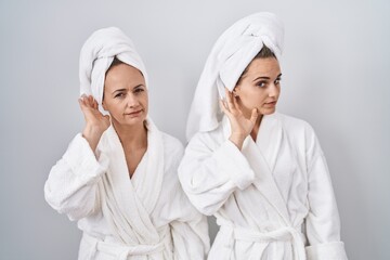 Canvas Print - Middle age woman and daughter wearing white bathrobe and towel smiling with hand over ear listening an hearing to rumor or gossip. deafness concept.