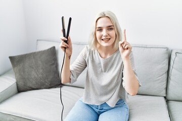 Wall Mural - Young caucasian woman at home holding hair straightener smiling with an idea or question pointing finger with happy face, number one