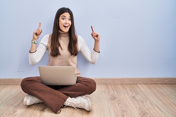 Sticker - Young brunette woman working using computer laptop sitting on the floor smiling amazed and surprised and pointing up with fingers and raised arms.