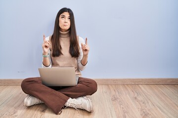 Poster - Young brunette woman working using computer laptop sitting on the floor pointing up looking sad and upset, indicating direction with fingers, unhappy and depressed.