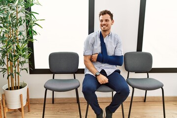 Canvas Print - Handsome young man sitting at doctor waiting room with arm injury looking away to side with smile on face, natural expression. laughing confident.