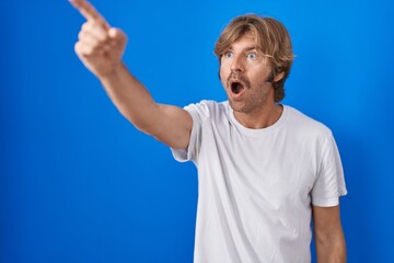 Poster - Middle age man standing over blue background pointing with finger surprised ahead, open mouth amazed expression, something on the front