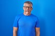 Hispanic man with grey hair standing over blue background sticking tongue out happy with funny expression. emotion concept.
