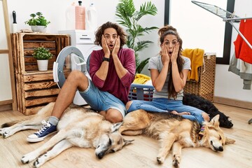 Poster - Young hispanic couple doing laundry with dogs afraid and shocked, surprise and amazed expression with hands on face