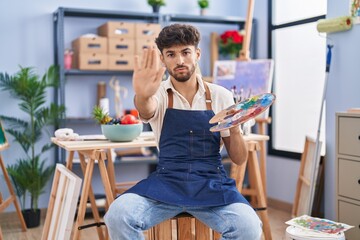 Sticker - Arab man with beard painter sitting at art studio holding palette with open hand doing stop sign with serious and confident expression, defense gesture