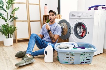 Wall Mural - Young hispanic man putting dirty laundry into washing machine surprised pointing with finger to the side, open mouth amazed expression.