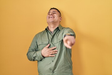 Poster - Hispanic young man standing over yellow background laughing at you, pointing finger to the camera with hand over body, shame expression