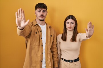 Poster - Young hispanic couple standing over yellow background doing stop sing with palm of the hand. warning expression with negative and serious gesture on the face.