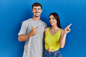 Poster - Young hispanic couple standing together over blue background cheerful with a smile on face pointing with hand and finger up to the side with happy and natural expression