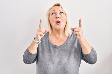 Sticker - Middle age caucasian woman standing over white background amazed and surprised looking up and pointing with fingers and raised arms.