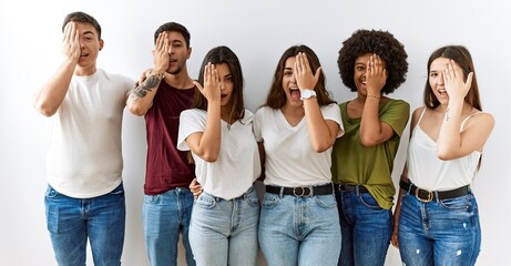 Poster - Group of young friends standing together over isolated background covering one eye with hand, confident smile on face and surprise emotion.