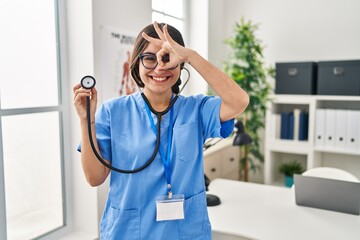 Poster - Young hispanic doctor woman wearing stethoscope at the clinic smiling happy doing ok sign with hand on eye looking through fingers