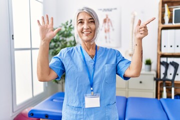 Poster - Middle age grey-haired woman wearing physiotherapist uniform at medical clinic showing and pointing up with fingers number seven while smiling confident and happy.