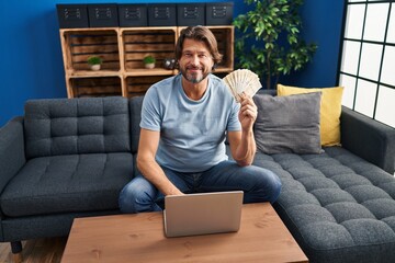 Canvas Print - Handsome middle age man holding 100 danish krone banknotes using laptop looking positive and happy standing and smiling with a confident smile showing teeth