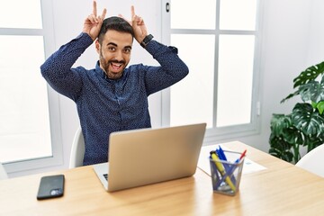 Sticker - Young hispanic man with beard working at the office with laptop posing funny and crazy with fingers on head as bunny ears, smiling cheerful