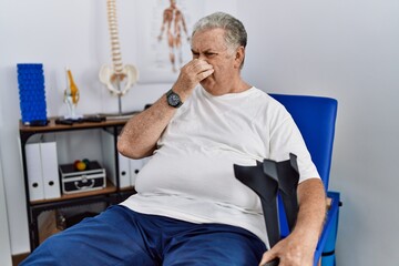 Wall Mural - Senior caucasian man at physiotherapy clinic holding crutches smelling something stinky and disgusting, intolerable smell, holding breath with fingers on nose. bad smell