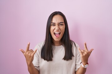 Wall Mural - Young hispanic woman standing over pink background shouting with crazy expression doing rock symbol with hands up. music star. heavy music concept.
