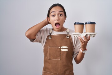 Sticker - Young hispanic woman wearing professional waitress apron holding coffee crazy and scared with hands on head, afraid and surprised of shock with open mouth