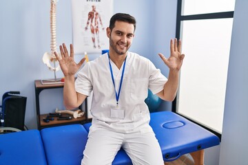 Sticker - Young hispanic man with beard working at pain recovery clinic showing and pointing up with fingers number nine while smiling confident and happy.