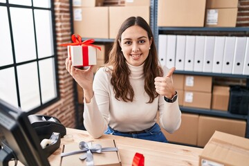 Sticker - Young hispanic doctor woman working at small business ecommerce holding present smiling happy and positive, thumb up doing excellent and approval sign