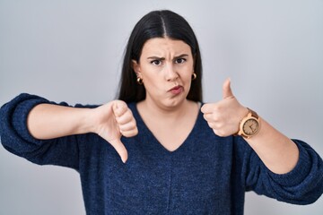 Poster - Young brunette woman standing over isolated background doing thumbs up and down, disagreement and agreement expression. crazy conflict