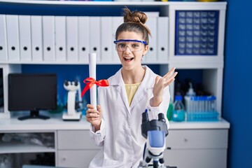 Sticker - Teenager girl working at scientist laboratory holding degree celebrating victory with happy smile and winner expression with raised hands