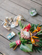 The bride's bouquet in a tropical style with exotic flowers lies on a wooden floor. Bouquet of protea, palm leaves, strelitzia, and red ginger. In the background are women's shoes.