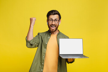 Yes. Overjoyed middle aged man holding pc laptop with blank screen and shaking clenched fists, mockup, yellow background