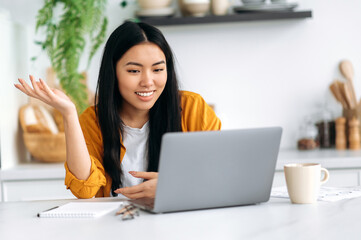 Online video communication. Friendly pretty asian brunette young woman, manager, freelancer, sit in the kitchen, talking on video call by laptop with the client or employees, discussing ideas, smiling