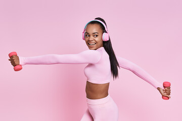 Wall Mural - Cheerful African woman in headphones exercising with dumbbells against pink background