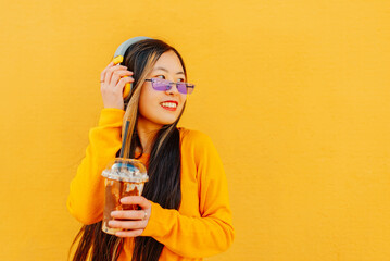 portrait of asian young girl listening to music with wireless headphones while drinking chocolate milkshake. Yellow background.