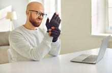Man With Rheumatoid Arthritis Kneads His Hands On Which He Wears Special Compression Gloves. Unhealthy Young Man With Sore Expression Feels Pain In His Hands While Working On Laptop. Health Concept.