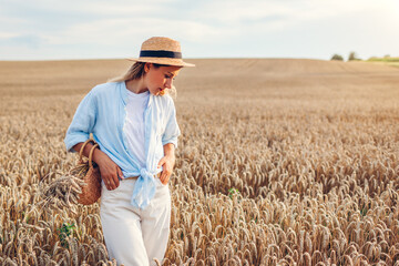 Wall Mural - Young woman walking in summer field wearing straw hat and linen shirt holding handbag with wheat bundle. Space