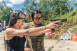 © PoppyPix - Male instructor showing female client how to aim handgun. Safety goggles and headphones. Firearms training at outdoor shooting range. Horizontal shot. High quality photo