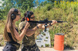 © PoppyPix - Woman standing next to man aiming submachine gun both wearing safety headphones and goggles. Firearms training at firing range. Outdoor horizontal shot. High quality photo
