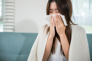 Ill Asian young woman cold covered with blanket sitting on sofa and sneeze with tissue paper at home, Female sick allergic blowing nose sneezing in tissue at home, Flu health care