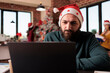 © DC Studio - Startup employee using laptop in business office to work on company project during winter season. Man with santa hat working on report in workplace with christmas tree and holiday decorations.