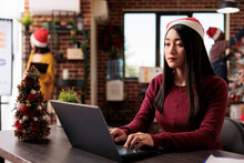 Asian Woman Working On Laptop In Decorated Office, Using Business Report To Work On Startup Company. Enjoying Christmas Tree And Festive Ornaments With Xmas Decorations At Job.