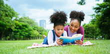Outdoor Of African American Little Girl Listen Mother Read A Book On The Grass. She Has A Look Of Pleasure And She Has Interested. African Happy Family Spend Time Together,woman Teaches At Park.