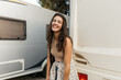 Positive caucasian young lady smiles broadly with teeth looking at camera relaxing at campsite. Brunette tourist wears undershirt, shirt and backpack. Happiness summer lifestyle concept.